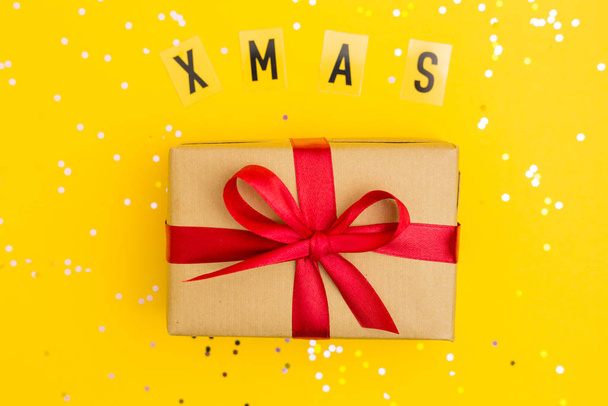 Bruine geschenkdozen met rode linten. Woord Kerstmis. Gele achtergrond met veelkleurige confetti. Vlakke lay stijl. Cadeau voor Kerstmis of Nieuwjaar - Foto, afbeelding