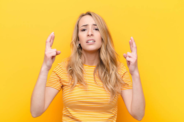 young pretty blonde woman crossing fingers anxiously and hoping for good luck with a worried look against flat color wall - Photo, image