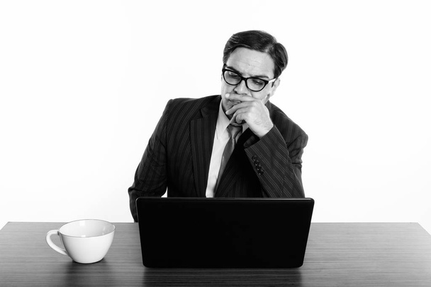 Hombre de negocios persa con anteojos mientras piensa y utiliza el ordenador portátil con taza de café en la mesa de madera
 - Foto, Imagen