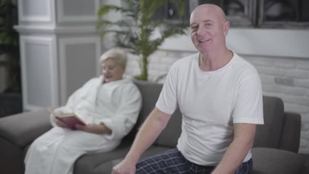 Happy mature Caucasian man looking back at wife, turning back and looking at camera. Blurred senior blond woman reading book at the background and waving to her husband. - Filmagem, Vídeo