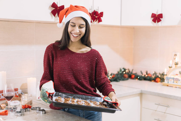 Happy young lady wearing Santa hat getting Christmas cookies out of the oven - Photo, Image
