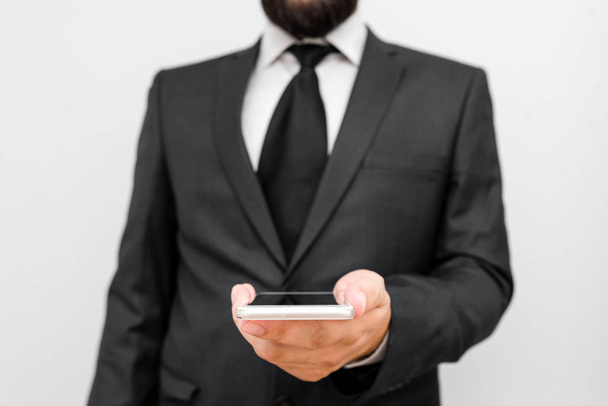 Мужчина с бородой носит официальную рабочую одежду с высокотехнологичным устройством смартфона. Мужчина в рабочем костюме плюс галстук с маленьким мобильным высокотехнологичным телефоном одной рукой - Фото, изображение