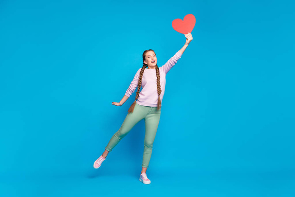 Full length photo of cheerful youth girl with braids pigtails hold hand catch flying paper card heart surprise on 14-february wear pink sweater green stylish pants isolated blue color background - Fotografie, Obrázek