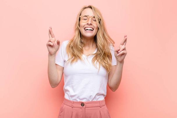 young pretty blonde woman smiling and anxiously crossing both fingers, feeling worried and wishing or hoping for good luck against flat color wall - Photo, Image