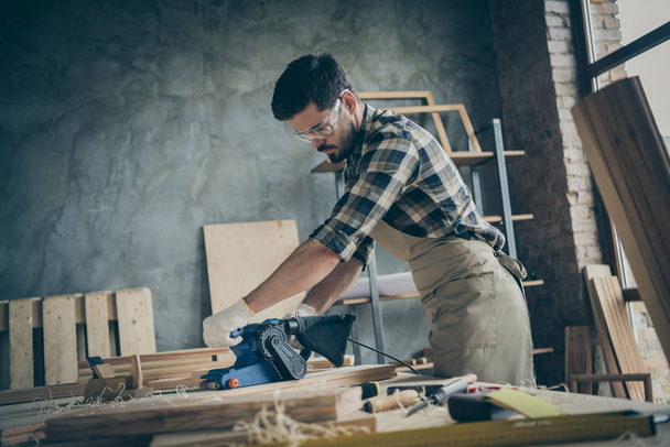 Side profile photo of serious pensive confident man processing wood with sander machine with eyes protected eye wear in checkered shirt gloves - Fotó, kép