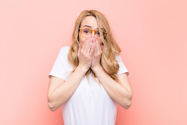 young pretty blonde woman happy and excited, surprised and amazed covering mouth with hands, giggling with a cute expression against flat color wall - Foto, immagini