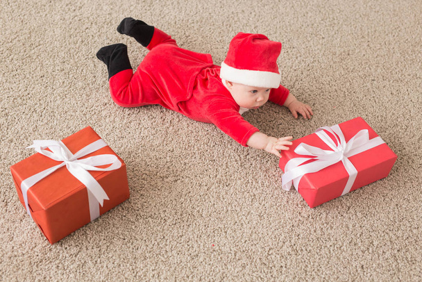 Beautiful little baby celebrates Christmas. New Years holidays. Baby in a Christmas costume and in santa hat and gift box, top view - 写真・画像