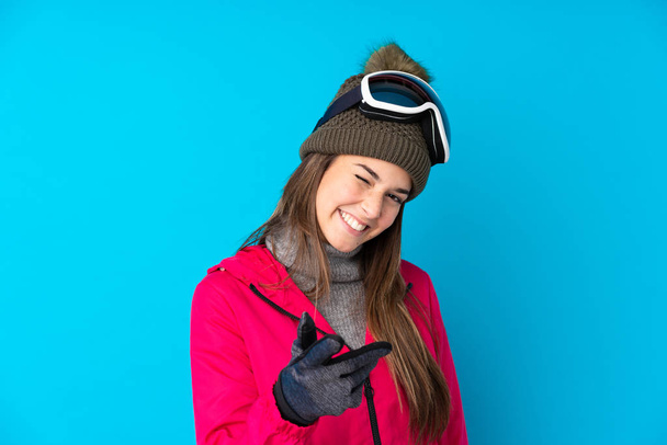 Adolescente skieuse avec des lunettes de snowboard sur fond bleu isolé pointe doigt à vous - Photo, image