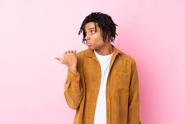 Jovem afro-americano com jaqueta de veludo sobre fundo rosa isolado infeliz e apontando para o lado
 - Foto, Imagem
