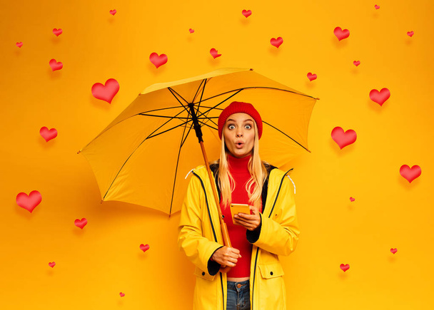 Blonde cute girl protects herself with umbrella due to rain of hearts on her smartphone. happy and surprised expression face. background - Фото, изображение
