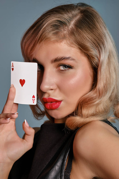 Blond meisje in zwart lederen jurk met aas van harten, poseren tegen grijze achtergrond. Gokken entertainment, poker, casino. Close-up. - Foto, afbeelding