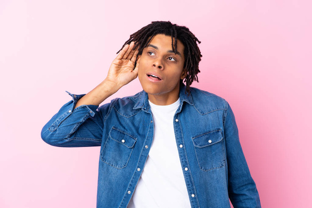 Молодий афроамериканець з джинсовою сорочкою через ізольоване рожеве тло слухає щось. - Фото, зображення