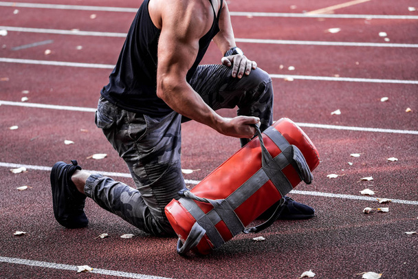 Strong male doing workout outdoor. Lifting heavy 10 kilo tool. Handsome sportsman. Young bodybuilder. Going sport. Sportsground background. Closeup - Photo, image