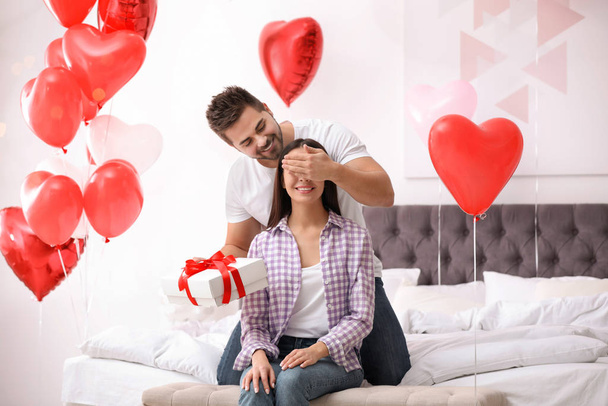 Young man presenting gift to his girlfriend in bedroom decorated with heart shaped balloons. Valentine's day celebration - Fotó, kép