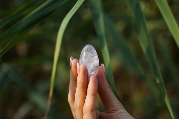 Female hand with french manicure holding transparent violet amethyst yoni egg for vumfit, imbuilding or meditation. Crystal quartz egg in hands on green background outdoors - Photo, Image