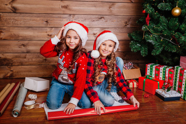 Two happy little girls in Santa hats pack gifts near Christmas tree - Фото, изображение