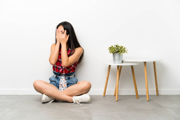 Подросток азиатская девушка сидит на полу покрывая глаза и глядя сквозь пальцы
 - Фото, изображение