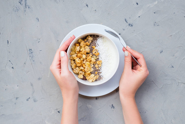 Schüssel mit hausgemachtem Müsli mit Joghurt und Chiasamen auf Steinhintergrund von oben gesehen. Frühstück. junge Frau beim Müsli essen in einem weißen Teller. Frauenhände halten Frühstücksschale - Foto, Bild