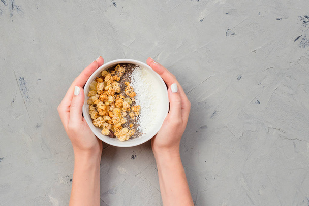 Чаша домашней мюсли с семенами йогурта и чиа на каменном фоне сверху. Пора завтракать. Молодая женщина ест мюсли в белой тарелке. Женские руки держат миску для завтрака
 - Фото, изображение
