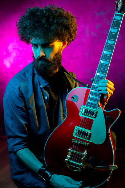 Close-up photo of hipster man with red guitar in neon lights. Rock musician is playing electrical guitar. - Fotó, kép