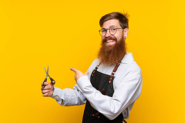 Barber with long beard in an apron over isolated yellow background and pointing it - Photo, Image
