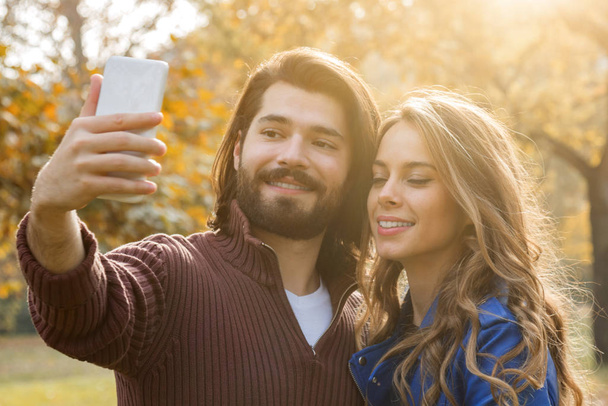 Νεαρό ζευγάρι χρησιμοποιώντας κινητό στο φθινόπωρο χρωματιστά πάρκο. - Φωτογραφία, εικόνα