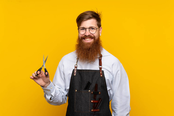 Barber with long beard in an apron over isolated yellow background smiling a lot - Photo, Image