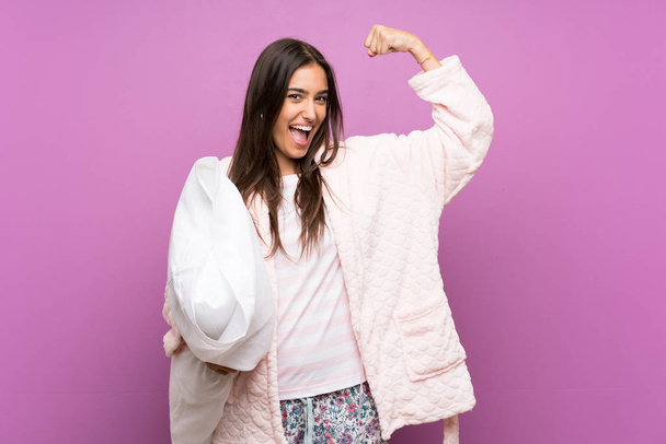 Young woman in pajamas and dressing gown over isolated purple background making strong gesture - Photo, Image