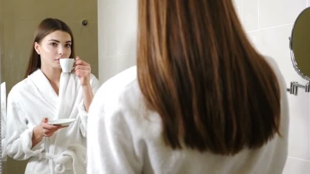 Giovane donna attraente in accappatoio bianco bere caffè davanti allo specchio del bagno. I filmati della vista posteriore. Bella donna guardando il suo riflesso nello specchio del bagno e sorridendo mentre si gode la mattina
 - Filmati, video