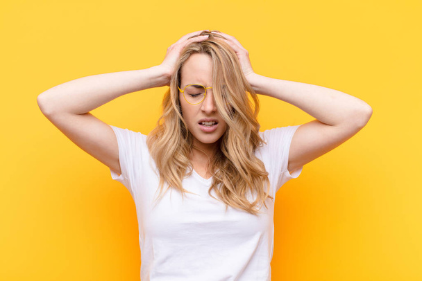 young pretty blonde woman feeling stressed and frustrated, raising hands to head, feeling tired, unhappy and with migraine against flat color wall - Фото, зображення