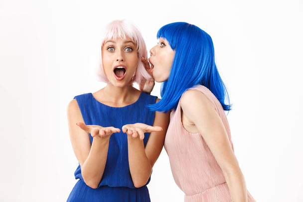 Portrait of two surprised women wearing blue and pink wigs talking secrets in ear while holding copyspace - Foto, imagen