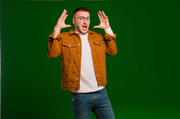 young handsome man screaming with hands up in the air, feeling furious, frustrated, stressed and upset against flat background - Photo, Image