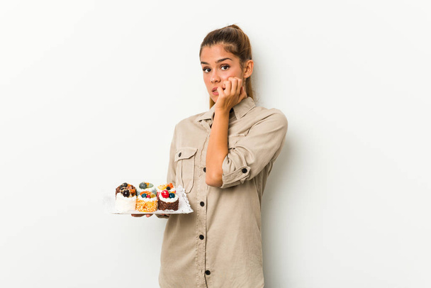 Mulher caucasiana jovem segurando bolos doces mordendo unhas, nervoso e muito ansioso
. - Foto, Imagem