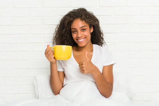 Nuori afrikkalainen amerikkalainen nainen istuu sängyllä pitäen kahvimukia hymyillen ja nostaen peukaloa
 - Valokuva, kuva