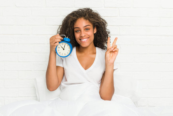 Молодая африканская американка, сидящая на кровати с будильником, показывает номер два пальцами
. - Фото, изображение
