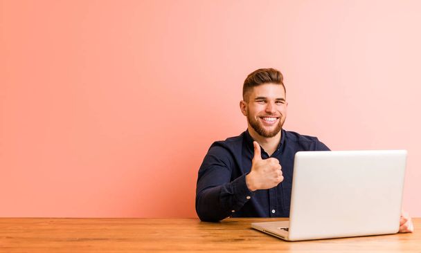 Nuori mies työskentelee kannettavan tietokoneen kanssa hymyillen ja nostaen peukalon ylös
 - Valokuva, kuva