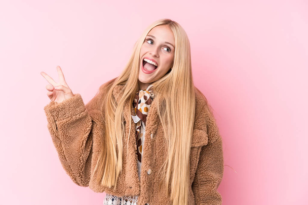 Young blonde woman wearing a coat against a pink background joyful and carefree showing a peace symbol with fingers. - Photo, Image