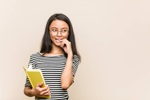 Mladá asijka studentka drží knihu uvolněně přemýšlet o něčem, co se dívá na kopii prostoru. - Fotografie, Obrázek