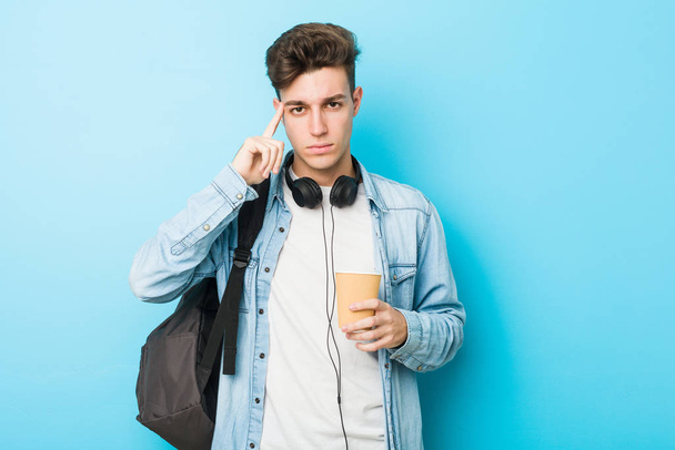 Jovem estudante caucasiano segurando um café take away apontando seu templo com o dedo, pensando, focado em uma tarefa
. - Foto, Imagem