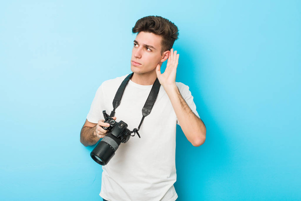 Νεαρός καυκάσιος φωτογράφος κρατάει κάμερα προσπαθώντας να ακούσει ένα κουτσομπολιό. - Φωτογραφία, εικόνα