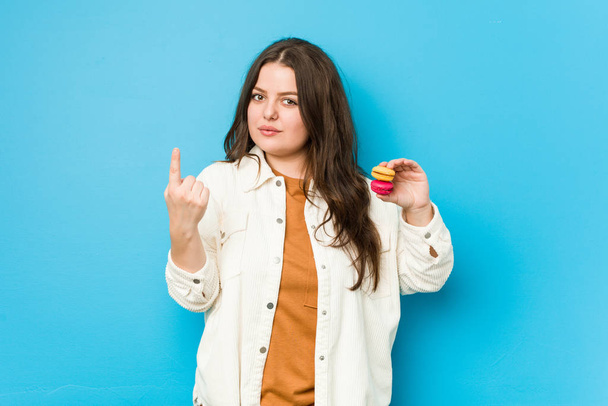 Молода пишна жінка тримає макаруни, вказуючи пальцем на вас, ніби запрошує наблизитися
. - Фото, зображення