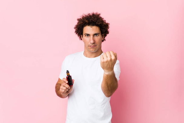Young caucasian curly man holding a vape showing fist to camera, aggressive facial expression. - 写真・画像