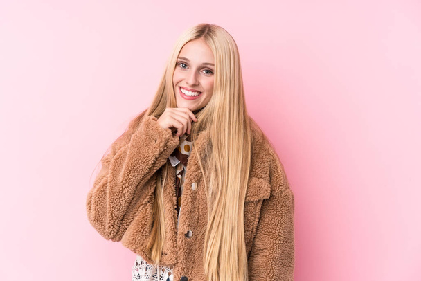 Young blonde woman wearing a coat against a pink background smiling happy and confident, touching chin with hand. - 写真・画像