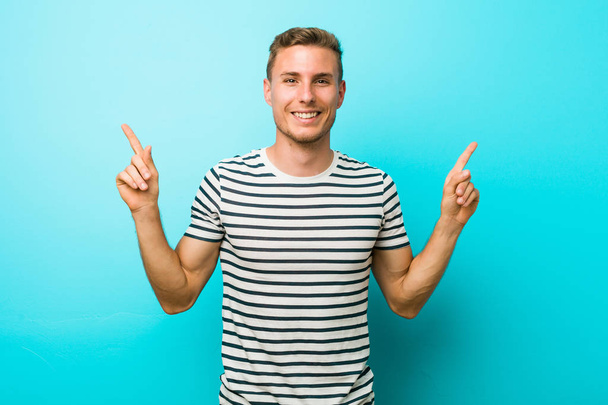 Giovane uomo caucasico contro un muro blu indica con entrambe le dita anteriori verso l'alto mostrando uno spazio vuoto
. - Foto, immagini