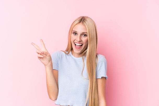 Mladá blondýna žena na růžovém pozadí radostné a bezstarostné ukazující symbol míru s prsty. - Fotografie, Obrázek