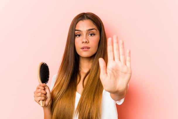 Mujer caucásica joven sosteniendo un cepillo de pelo de pie con la mano extendida mostrando señal de stop, lo que le impide
. - Foto, imagen