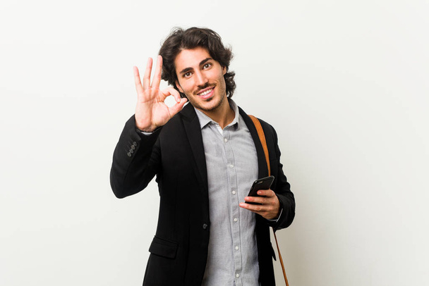 Joven hombre de negocios sosteniendo un teléfono alegre y confiado mostrando gesto ok
. - Foto, Imagen