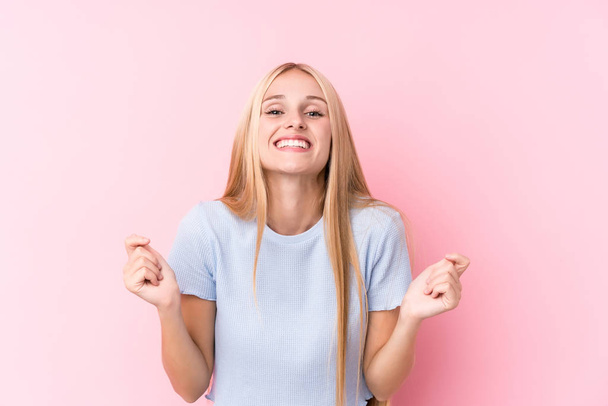 Young blonde woman on pink background raising fist, feeling happy and successful. Victory concept. - Photo, Image