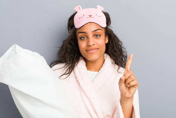 Młoda Afroamerykanka w piżamie i masce do spania trzymająca poduszkę z palcem numer jeden. - Zdjęcie, obraz