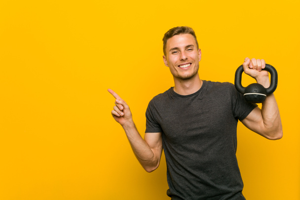 Young caucasian man holding a dumbbell smiling cheerfully pointing with forefinger away. - Photo, Image